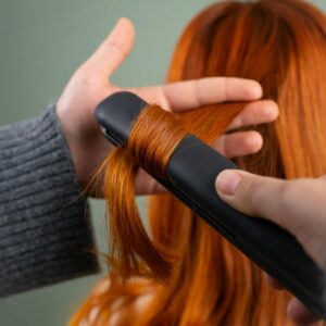 Close-up of red hair being styled with a flat iron indoors.